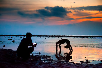 The girl shows the figures standing in the bridge, on hands with a bent back, the guy shoots video on the smartphone, against the sunset at the sea. Gili Trawangan island, Lombok, Indonesia.