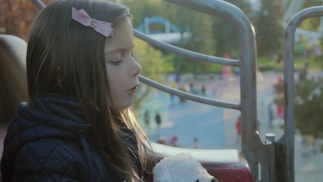 Happy little girl with teddy bear in amusement park on carousel.