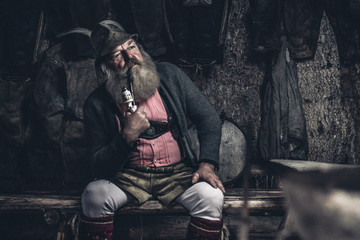 thoughtful old full bearded man with traditional austrian garb sitting inside a wooden cabin smoking a pipe