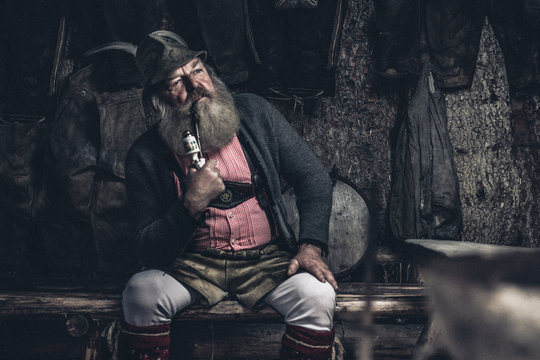 thoughtful old full bearded man with traditional austrian garb sitting inside a wooden cabin smoking a pipe