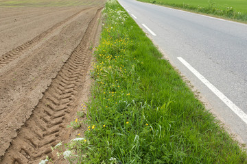 A brown field with ploughed rows of dirt with tractor tracks, next to grass and asphalt road