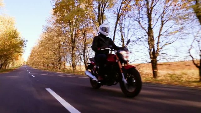Steadycam shot of motorcyclist driving his motorbike on the road during autumn sunset. Yellowleaves trees along the road
