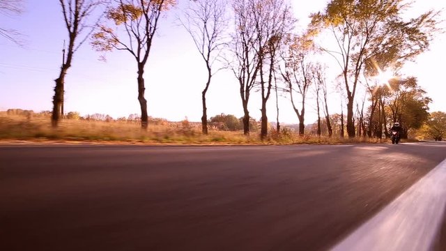 Steadycam shot of motorcyclist driving his motorbike on the road during autumn sunset. Yellowleaves trees along the road