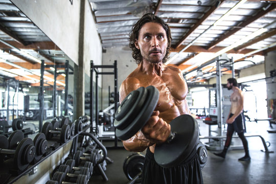 Ripped chiseled buff male bodybuilder flexing while curling dumbbell at gritty extreme gym