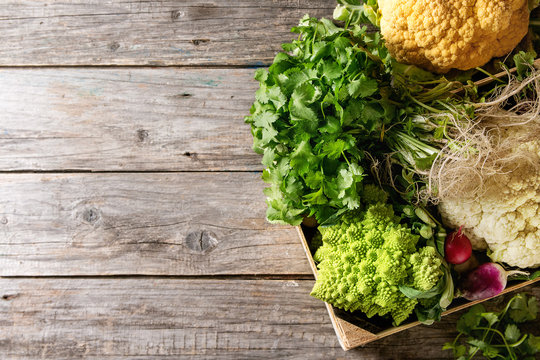 Variety of fresh raw organic colorful cauliflower, cabbage romanesco and radish with bundle of coriander in wood box over old wooden background. Top view with copy space. Food farm market concept