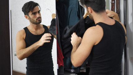 Sexy handsome man standing in his bedroom, dressing with shirt, next to mirror wardrobe door