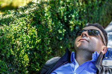 A man in a shirt and black glasses rests placidly on a bench