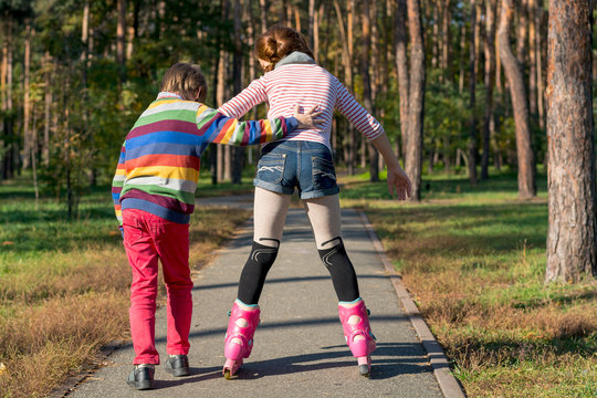 The boy helps the girl to roller-skate in the park. Brother supports his sister on the rollers.