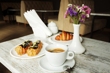White plate with Coffee eclairs and croissant with cup of fresh black coffee on wooden table. Top view.