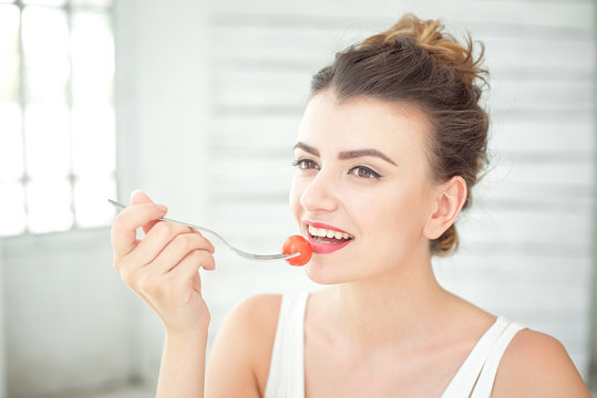Close up portrait of attractive healthy young woman eating green salad indoors holding fork with fresh cherry tomato.