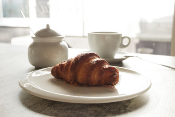White plate with croissant with cup of fresh black coffee on wooden table. Top view.