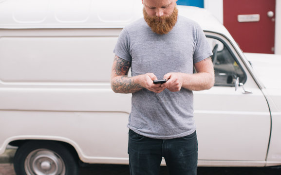 Bearded young man is texting on his cell phone with car on background