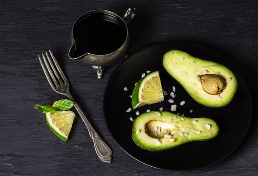 Avocados halves on black plate placed with lime slices,salt and balsamic vinegar on dark wooden background.Top view with copy space 