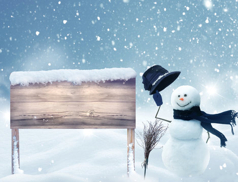 Merry Christmas and happy New Year greeting card with wooden signboard in snow. Snowman standing in winter Christmas landscape.Winter background