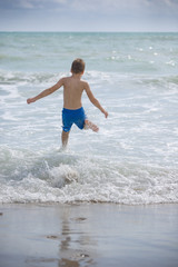 Fototapeta premium Back view on happy running kid boy rushing into ocean water on the beach. Child enjoying waves on the beach. Lifestyle