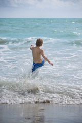 Back view on happy kid boy running into ocean or sea waves for swimming. Happy summer vacations.