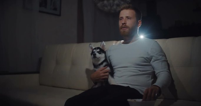 Young man with dog sitting on a sofa, watching drama on the TV at home in the living room and start cry.