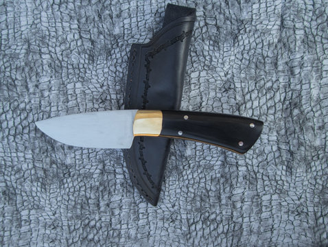 A fixed blade knife with a buffalo horn handle is displayed with its accompanying leather sheath on top of a black patterned fabric.