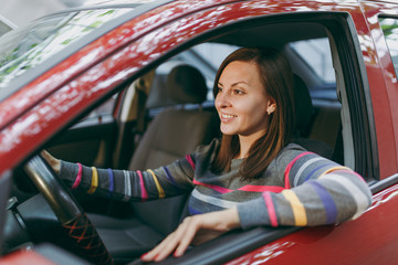 Beautiful young happy smiling European brown-haired woman with healthy clean skin dressed in a striped t-shirt sits in her red car with black interior. Traveling and driving concept.
