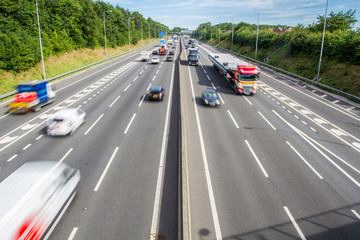 An aerial photograph of a busy motorway in the UK with three lanes and a junction and traffic including cars and trucks speeding by with blurred motion. - obrazy, fototapety, plakaty