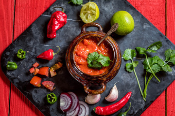 Homemade salsa with ingredients on black stone desk on red rustic background, tomato, pepper, onion, chili, habanero, coriander leaf. Mexican food concept