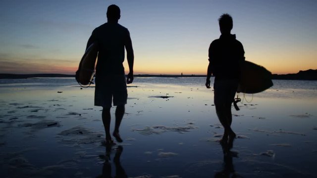 Silhouette of 2 surfers walking on beautiful sandy beach at sunset