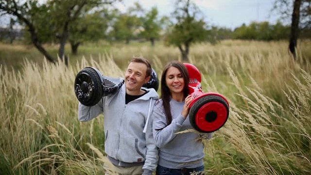Young man and girl go into the field with hoverboards look at one another in urban clothing. Steadicam