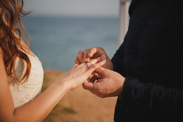 Man is putting wedding ring on his bride's hand, close up. Outdoor beach wedding ceremony, stylish...