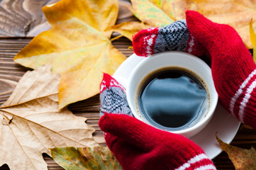 Hands in red knitted gloves holding a hot cup of coffee against yellow leaves background. Concept...