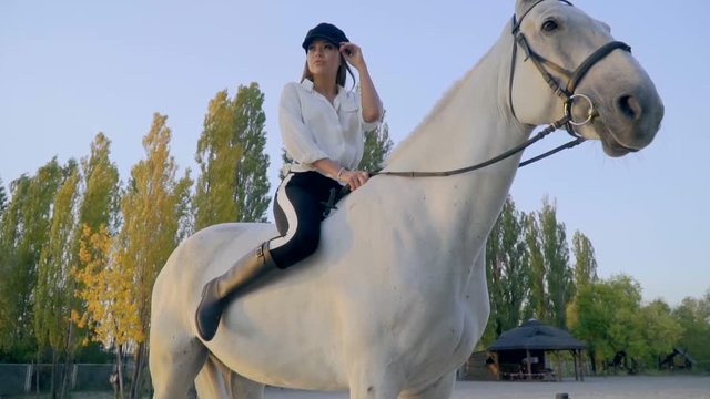 A female horse rider sits riding a white horse and looks into the distance adjusting her black cap, slow motion