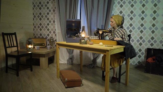 Retro seamstress girl sews cloth with old manual hand sewing machine. Woman working at home or workshop at night with kerosene lamp, listens music on gramophone or phonograph with vinyl plate