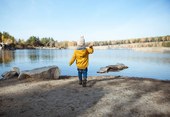 A little boy in a yellow jacket walks near the lake.