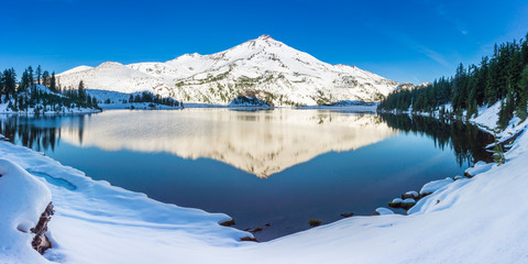 A snow covered mountain peak is reflected in the calm morning waters of a high elevation lake.