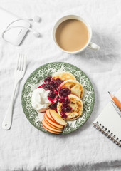 Cottage cheese pancakes with greek yogurt and cranberry sauce, coffee with milk, notebook, phone with headphones on white table, top view. Cozy morning breakfast