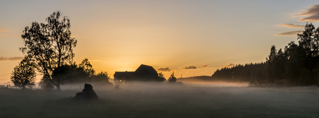Picture of barn with the sun behind it creating a yellowish glow with fog in the front. Photo taken in Sweden. 