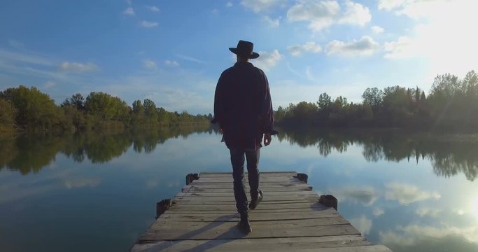 Giovane ragazzo con barba e cappello sta camminando su un molo di legno in riva a un lago 