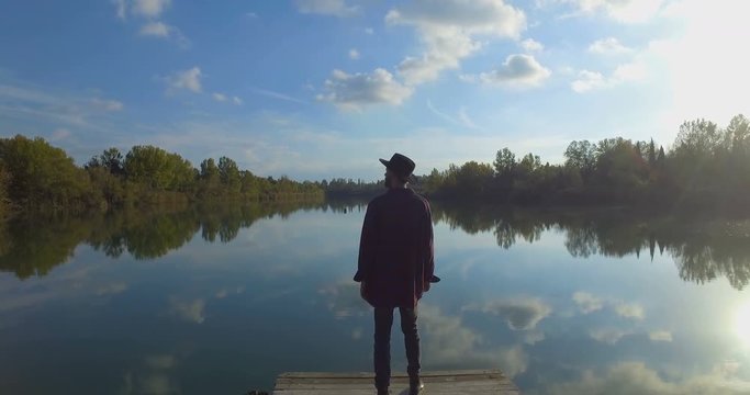 Giovane ragazzo con barba e cappello sta camminando su un molo di legno in riva a un lago 
