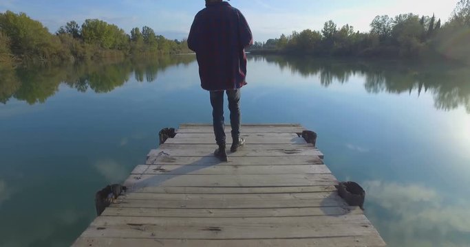 Giovane ragazzo con barba e cappello sta camminando su un molo di legno in riva a un lago 