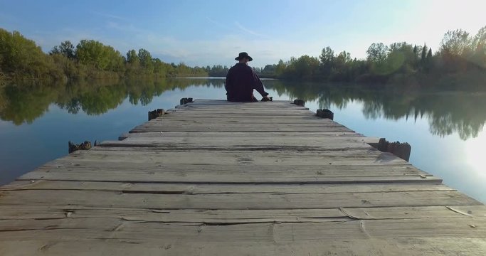Giovane ragazzo con barba e cappello sta camminando su un molo di legno in riva a un lago 