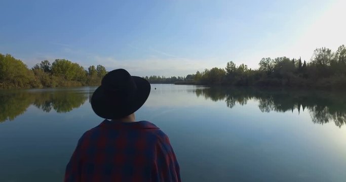 Giovane ragazzo con barba e cappello sta camminando su un molo di legno in riva a un lago 