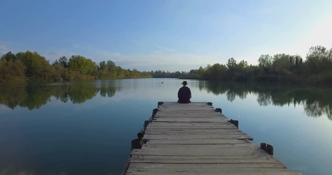 Giovane ragazzo con barba e cappello sta camminando su un molo di legno in riva a un lago 