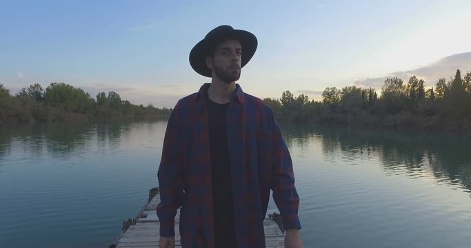 Giovane ragazzo con barba e cappello sta camminando su un molo di legno in riva a un lago 