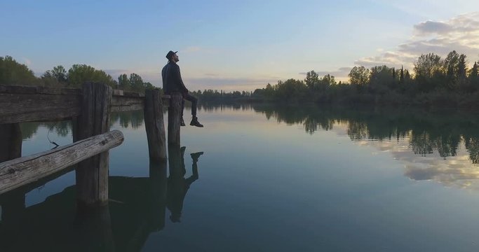 ragazzo con il cappello seduto su un molo di legno guarda tranquillo il lago blu al tramonto