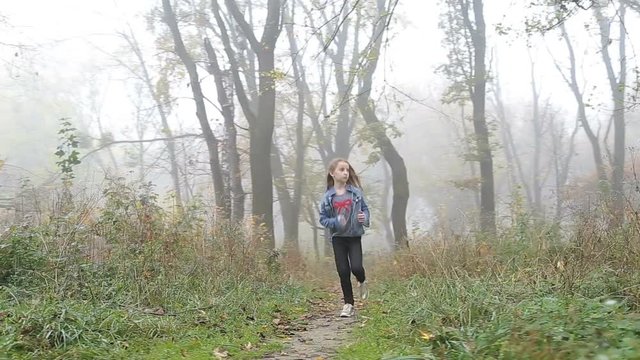 Little European girl with a long hair, blue jacket, black pants, sneakers and blue eyes. A frightened little child is running through the foggy deserted forest. Loneliness. Steady cam front shot.