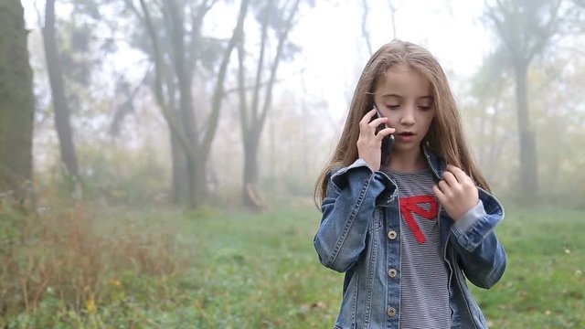 Little European girl with a long hair, blue jacket, black pants, sneakers and blue eyes. A frightened little child is walking through the foggy deserted forest and talking cell phone.  