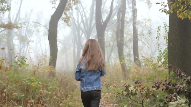 Little European girl with a long hair, blue jacket, black pants, sneakers and blue eyes. A scarried little child is running away through the foggy deserted forest. Loneliness. . Steady cam back shot.