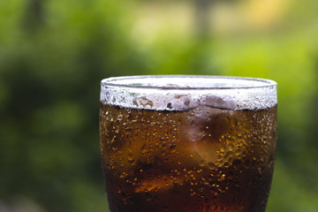 Glass of cola with ice cubes on table with green nature background. It is suitable for seasoning...