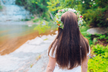 Beautiful young girl in a white chemise and a wreath of flowers on her head sitting stay near mountain river. 