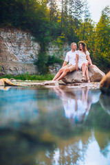 Couple in forest by the stream. Young couple walking on the rocks by the mountain stream with man helping his girlfriend. Funny moment with water. Man and woman sitting on stone.