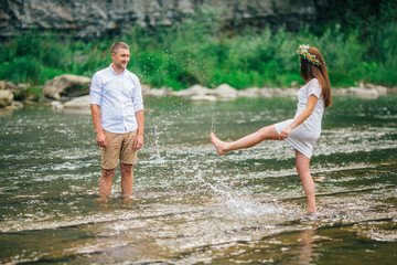 Couple in forest by the stream. Young couple walking on the rocks by the mountain stream with man helping his girlfriend. Funny moment with water. Man and woman sitting on stone.